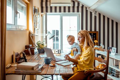 Een vrouw die aan een bureau zit en een baby vasthoudt en naar een laptop kijkt