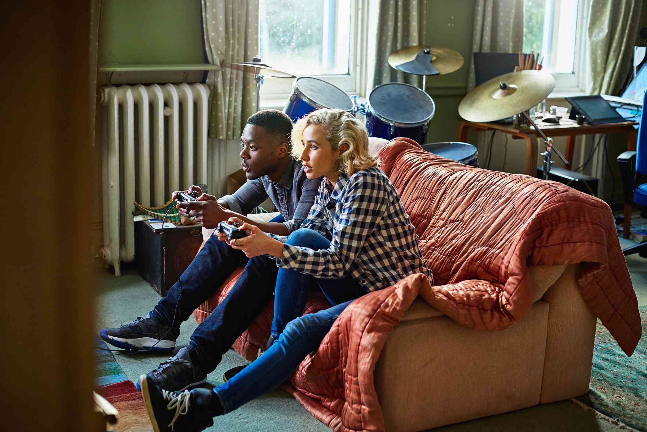 Twee jonge volwassenen die console-videogames spelen, zittend op een bank in een klein appartement.