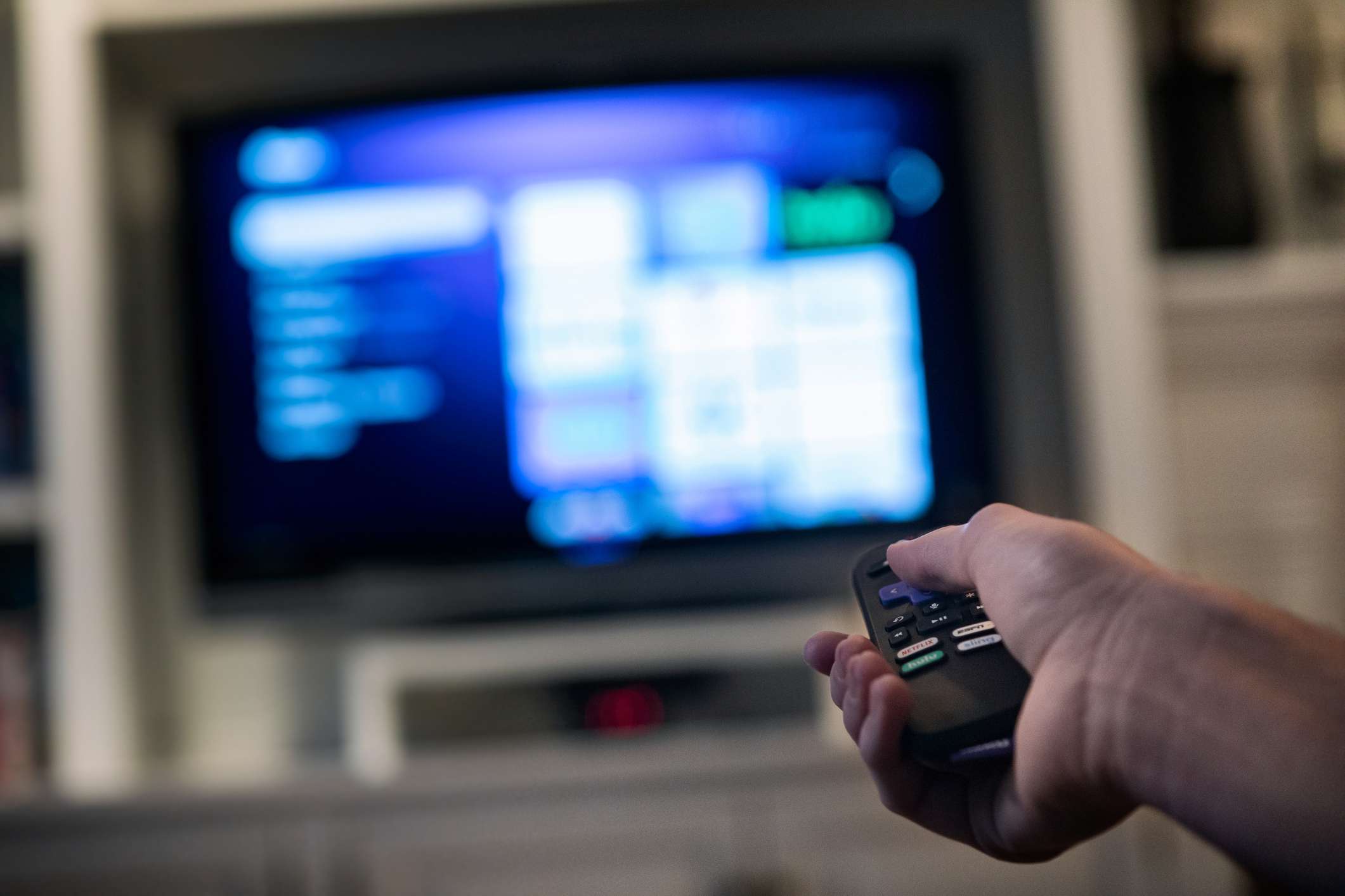 close-up van een hand die de afstandsbediening van een televisie vasthoudt met de tv op de achtergrond. 