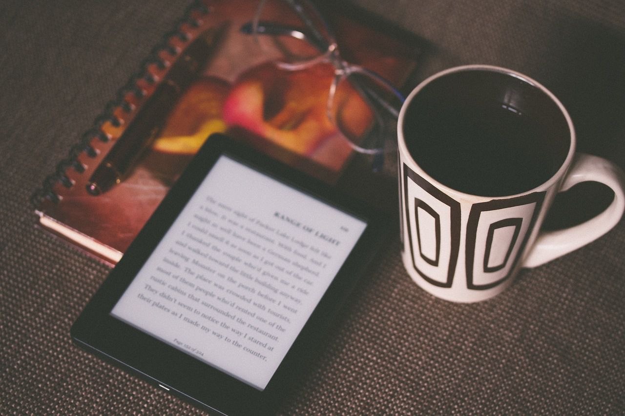 Afbeelding van een e-boek, een drankje, een notitieboekje en een bril op een tafel