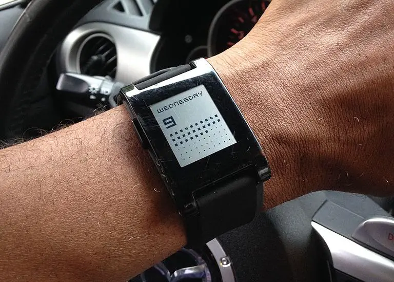 Een close-up van een Pebble-horloge om iemands pols met autodisplays op de achtergrond. 
