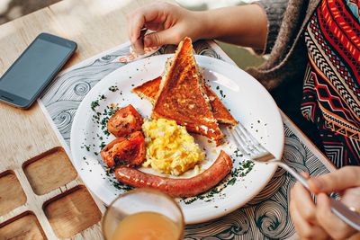 Close up van iemand die een ontbijtschotel eet: toast, eieren, tomaten en worst op een bord.  Op de tafel, naast het bord, ligt een openstaande, uitgeschakelde smartphone.