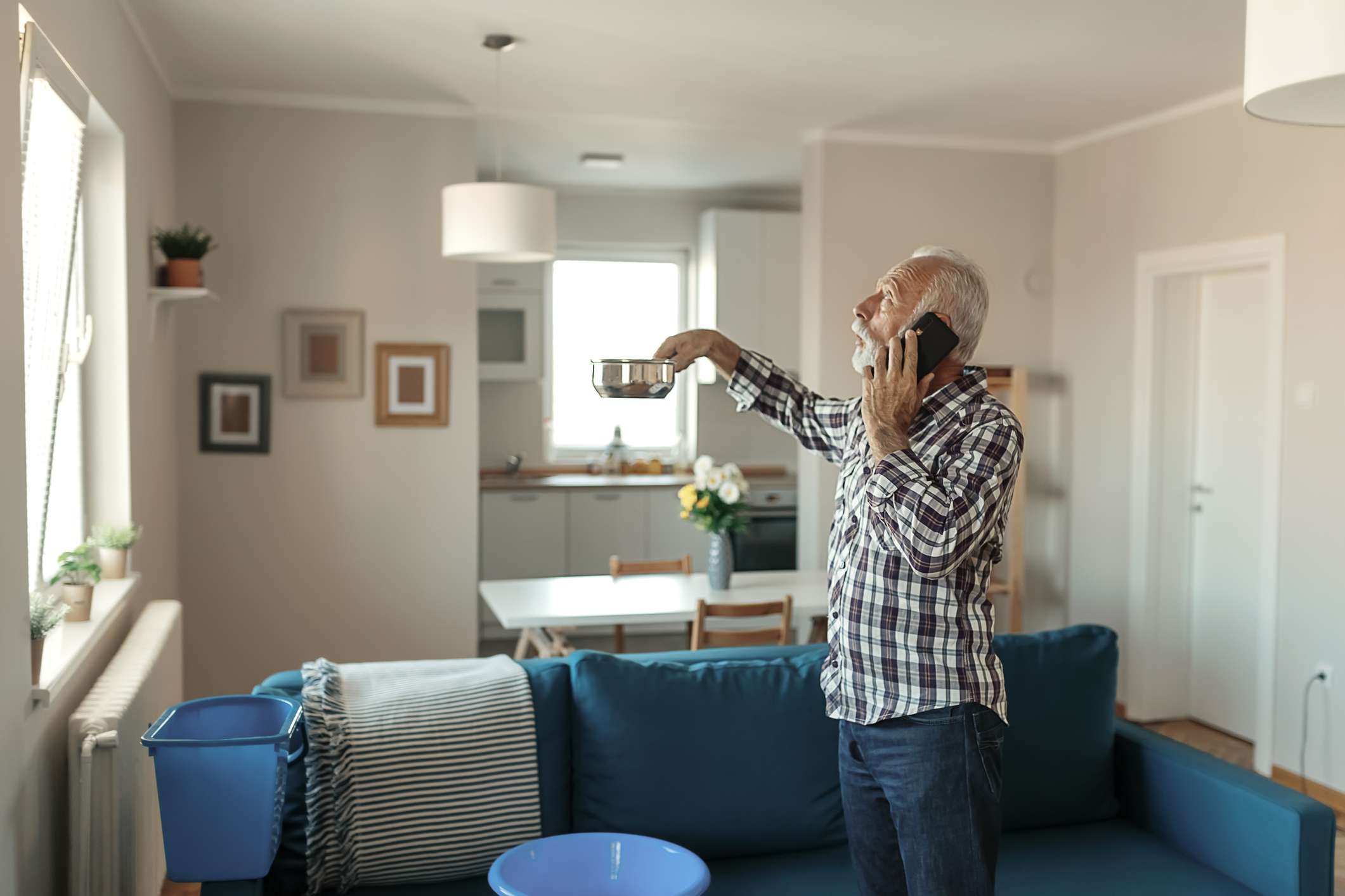 Bebaarde senior man praten op smartphone in de buurt van plastic waskom thuis in de woonkamer vanwege daklekkage