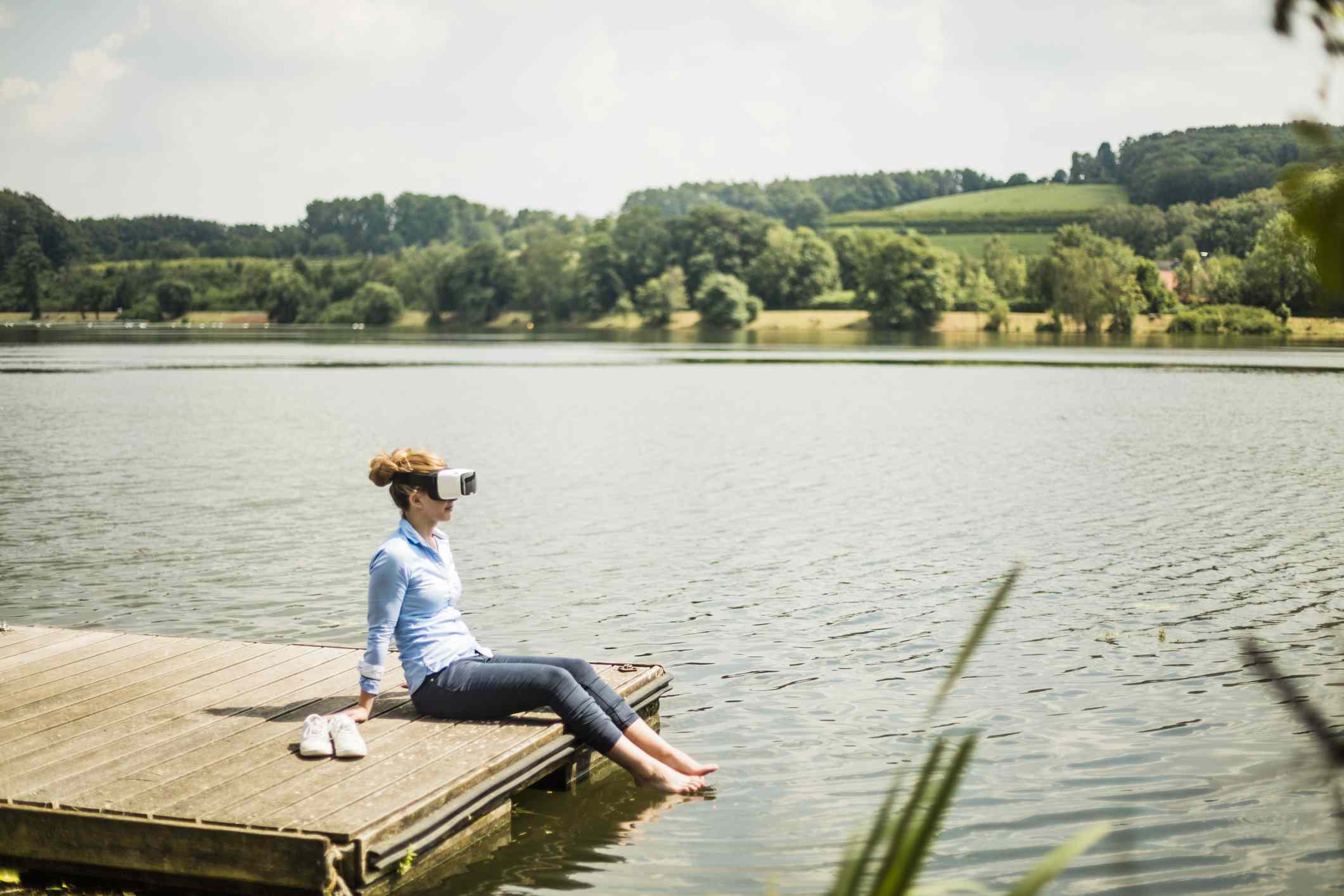 Iemand met een VR-bril zit op een steiger aan het meer met de voeten in het water. 