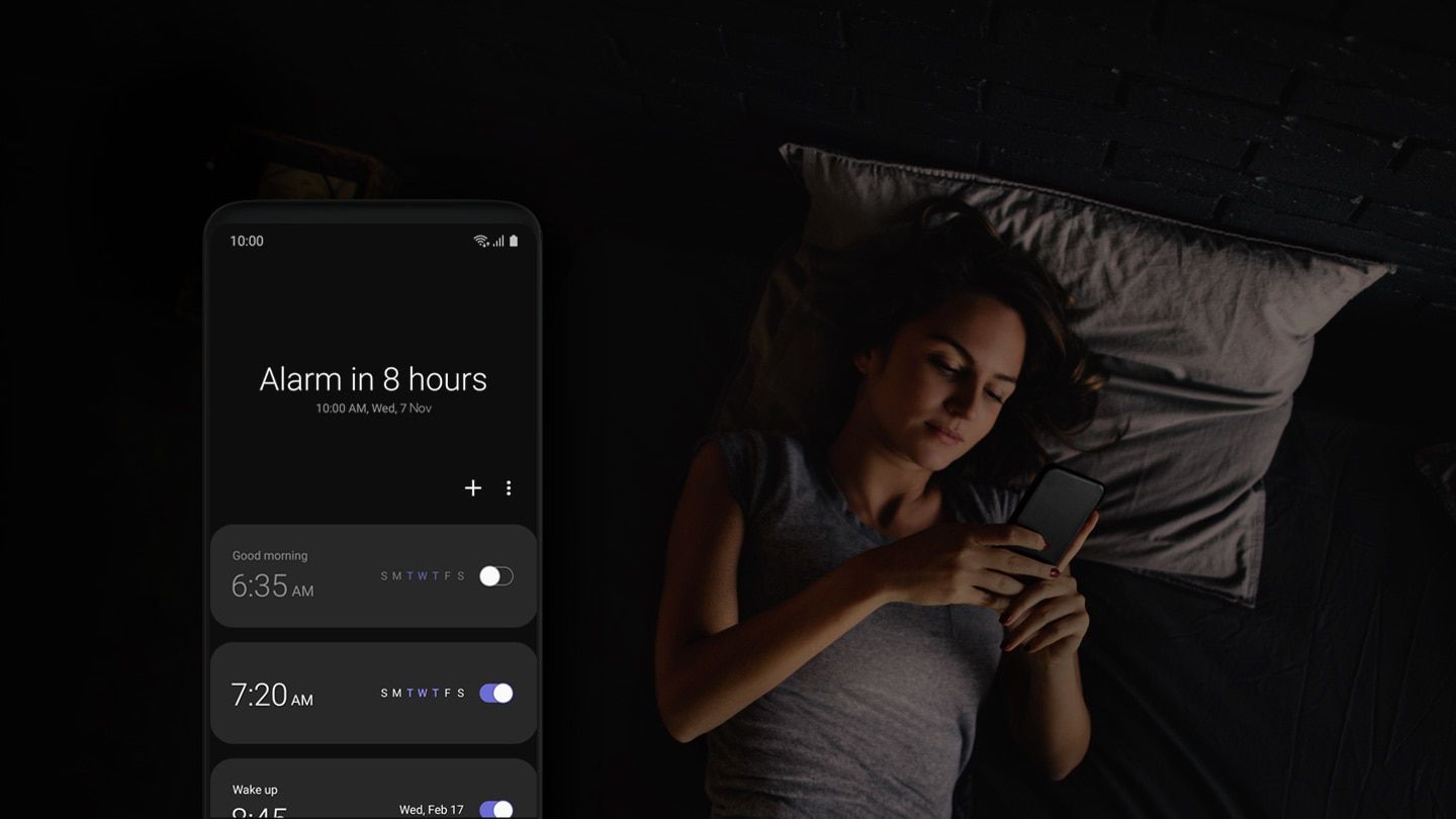 Vrouw in bed die haar telefoon gebruikt, alarminterface van One UI aan de linkerkant geplaatst