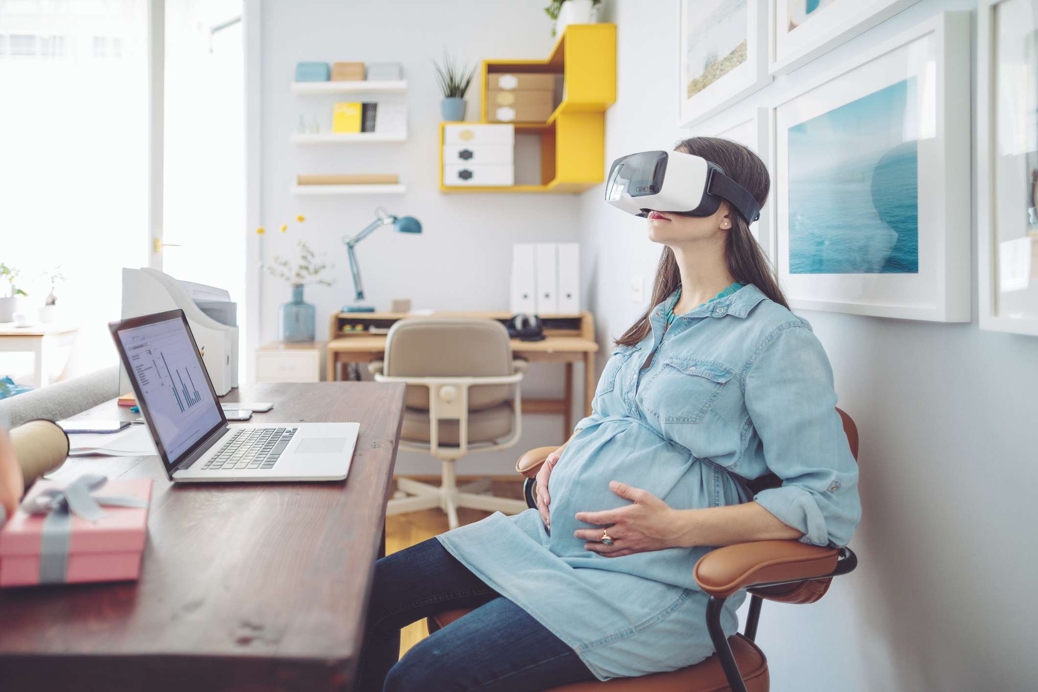 Een zwangere vrouw die een VR-headset draagt ​​terwijl ze in een thuiskantoor werkt met een laptop in de buurt.