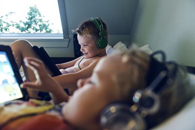Twee kinderen die op een bed liggen met game-apparaten in hun handen en een koptelefoon op