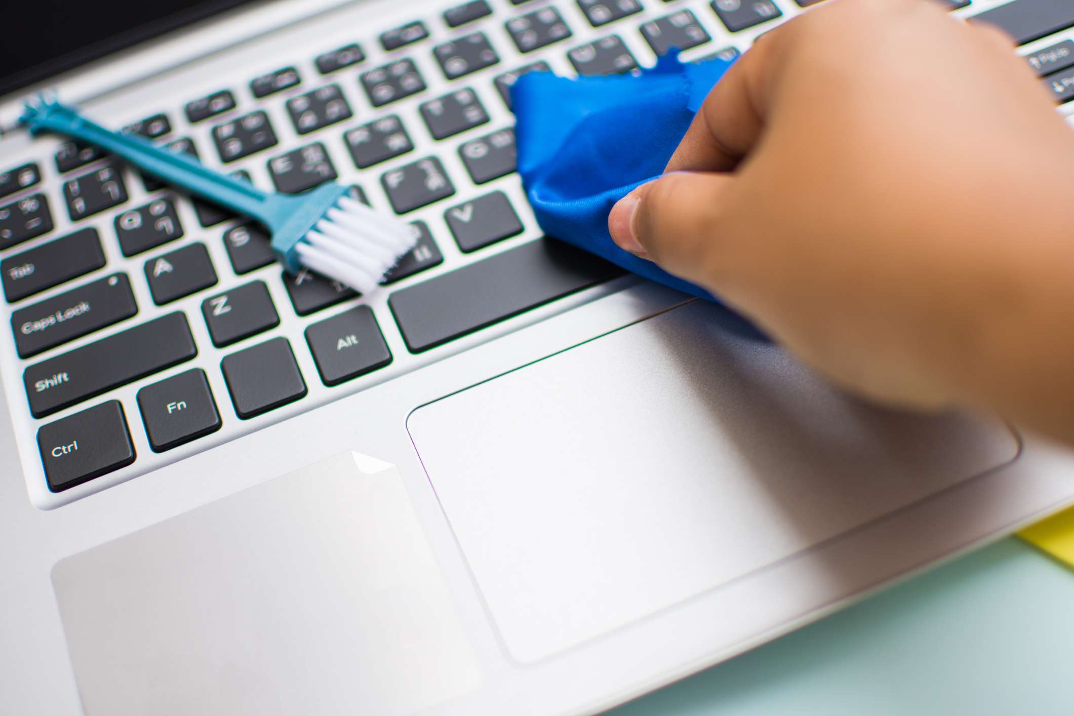 Een persoon die een blauwe doek over een laptoptoetsenbord veegt met een kleine borstel in de buurt