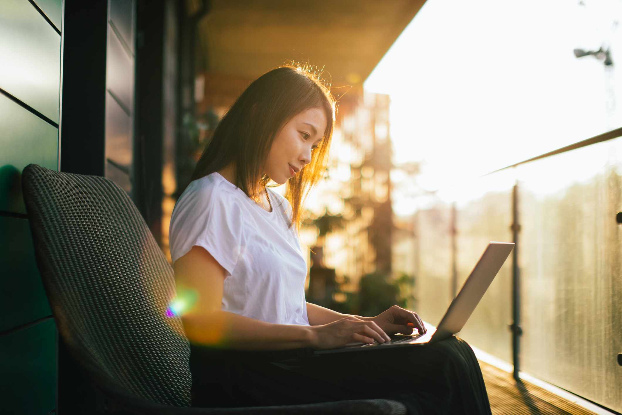 Vrouw die met de computer op haar schoot werkt, zittend op het balkon bij zonsondergang sunset