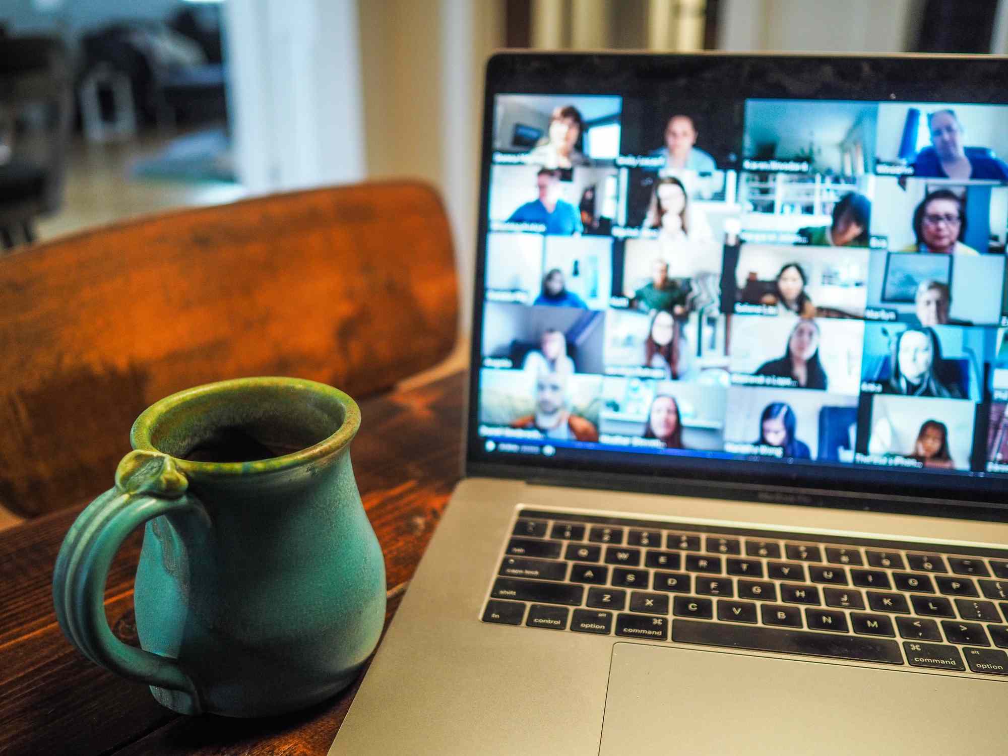 Een opengeklapte laptop op een keukentafel met een videogesprek voor meerdere personen met een kopje koffie in de buurt. 